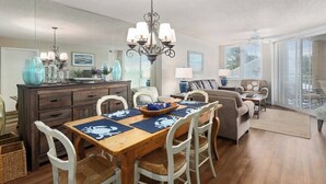 A dining area with a wooden table, white chairs, and a chandelier. There is a sofa and a lamp in the background. - Oceanfront Condominium! (Saint Simons Island)