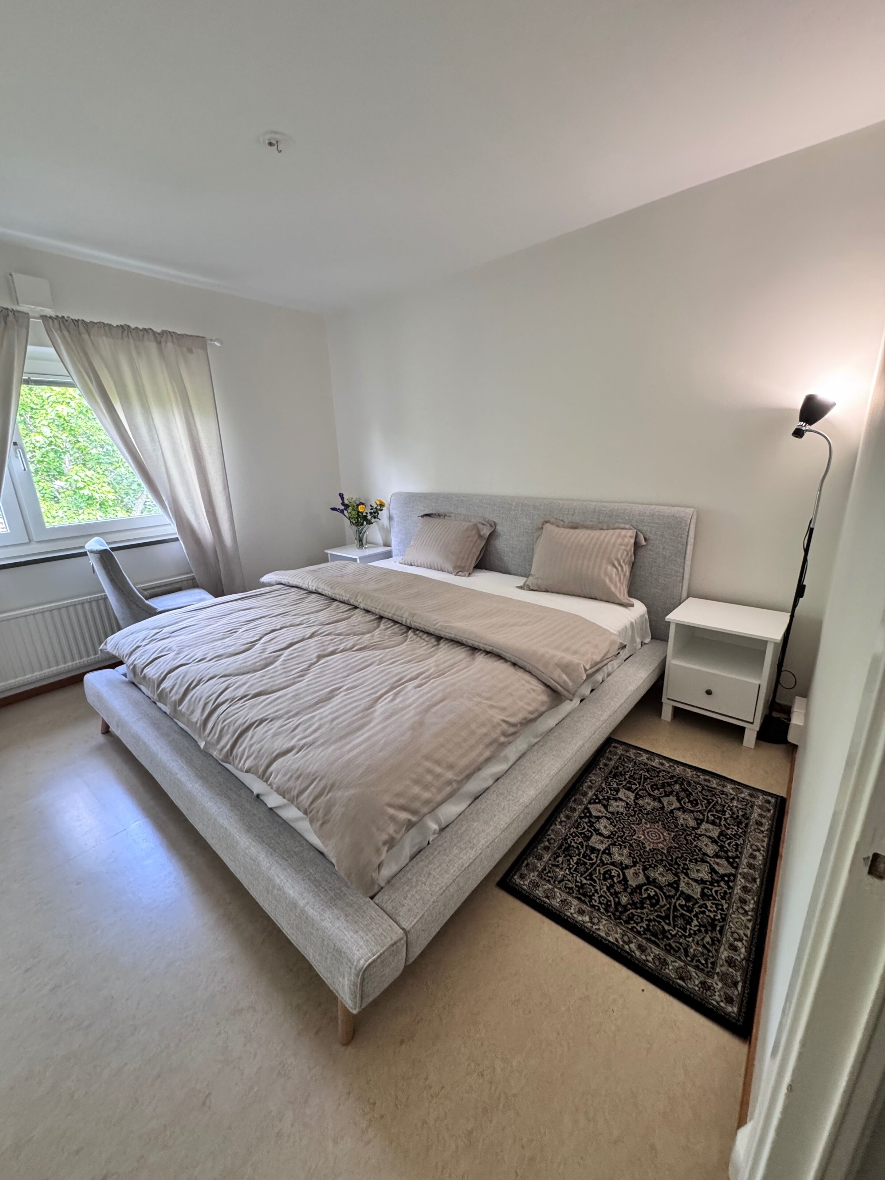 Une chambre à coucher comprenant un lit, des tables de chevet, une commode, une lampe de sol et une fenêtre avec des rideaux.