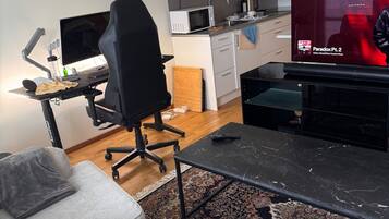 Un salon moderne avec un fauteuil de jeu noir, un canapé gris, une table basse noire et un téléviseur diffusant « Paradox Pt. 2 ». On aperçoit une cuisine avec un four à micro-ondes et des placards à l’arrière-plan.
