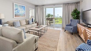 Una sala de estar con sofá, mesa de centro y televisor, ventilador de techo y vista a un paisaje verde a través de un ventanal.