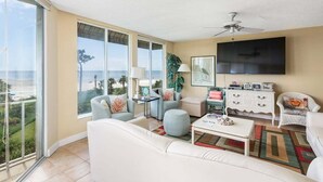A spacious living room with a white sofa, armchairs, a coffee table, and a TV. Large windows offer a view of the ocean. - Oceanfront Condominium with Gorgeous Ocean Views. (Saint Simons Island)