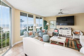 A spacious living room with a white sofa, armchairs, a coffee table, and a TV. Large windows offer a view of the ocean.