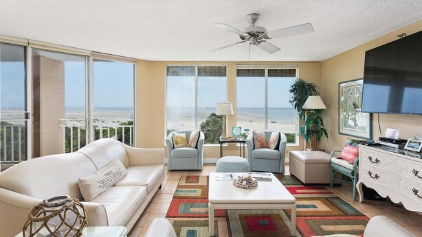 A spacious living room with a balcony overlooking the beach, featuring a white sofa, a coffee table, and a TV. - Oceanfront Condominium with Gorgeous Ocean Views. (Saint Simons Island)