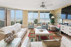 A spacious living room with a balcony overlooking the beach, featuring a white sofa, a coffee table, and a TV.