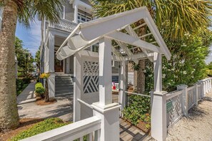 A white wooden deck with a pergola, surrounded by palm trees and a pathway.
