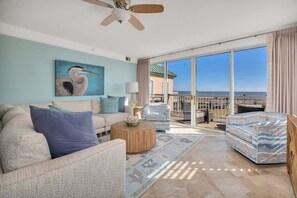 A spacious living room with a large window, a ceiling fan, a painting of a bird, and a view of the ocean.