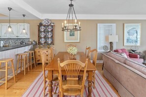 A dining area with a wooden table, chairs, and a chandelier. There is a kitchen with white cabinets and a sink. A sofa and a lamp are visible in the background. - Enjoy Island Living at Beachfront Condominium! (Saint Simons Island)