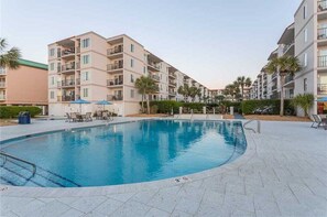 A swimming pool surrounded by multi-story buildings with balconies, a clear sky, and a few palm trees.