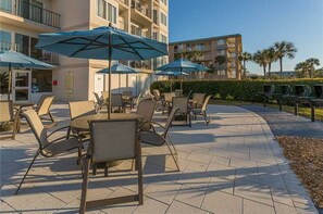 An outdoor patio with tables and chairs, umbrellas, and a building in the background. - Beachfront Condominium Perfect for Small Families! Pool, Fitness Center, Tennis! (Saint Simons Island)