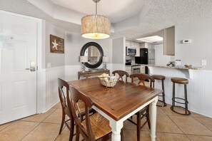 A dining area with a wooden table, chairs, and a chandelier. There is a kitchen in the background with appliances and a counter. - Oceanfront Condominium with Great Amenities (Saint Simons Island)