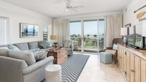 Ein geräumiges Wohnzimmer mit grauem Sofa, einem Holztisch und einem Fernseher auf einer Holzkommode. Es gibt ein großes Fenster mit Blick ins Freie.