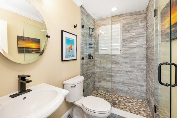 A bathroom with a white sink, toilet, and shower. There is a glass shower enclosure with a wooden-textured wall and a framed poster on the wall.