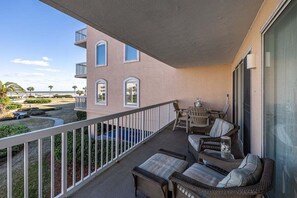 Un balcon agrémenté de meubles en osier, une vue sur la plage et un immeuble à la façade rose.