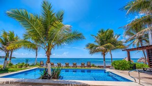 A pool area with lounge chairs and palm trees, overlooking a beach and ocean. - Large upscale penthouse, just right for families or friends, Pool, AC & WiFi! (Akumal)