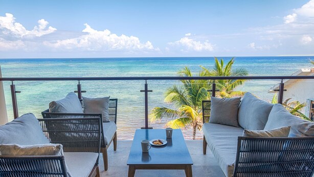 A balcony with wicker furniture, a table with a plate and mug, and a view of the ocean. - Large upscale penthouse, just right for families or friends, Pool, AC & WiFi! (Akumal)