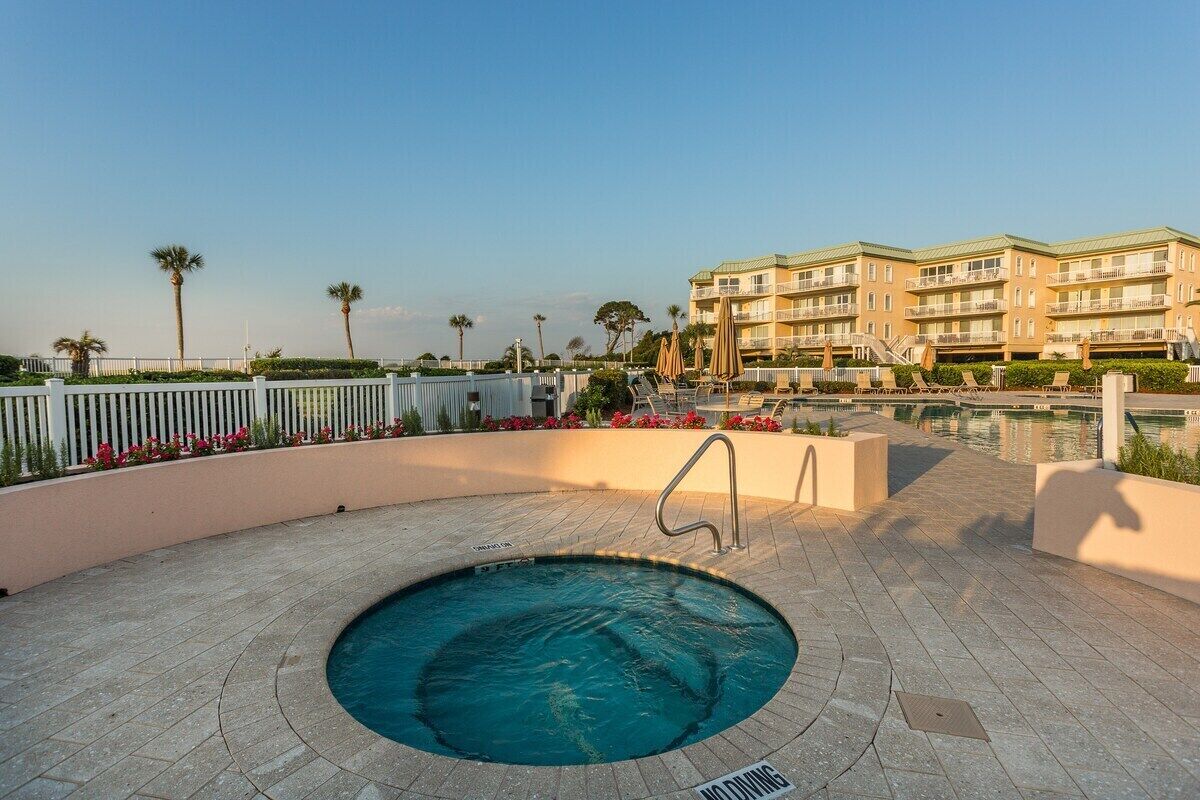 A swimming pool area with a hot tub, surrounded by a white fence, palm trees, and a building with balconies in the background.