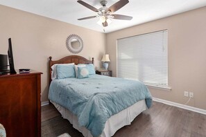 A bedroom with a bed, a ceiling fan, a bedside table with a lamp, a TV on a stand, and a window with blinds.