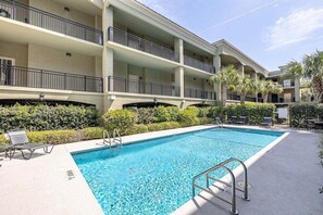 A swimming pool with a metal ladder, surrounded by a multi-story building with balconies and palm trees.