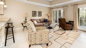 A spacious living room with a sofa, armchair, and a glass coffee table. There is a ceiling fan, a small table with a lamp, and a view of the outdoors through large windows. - Half a Mile to the Pier and Village! (Saint Simons Island)