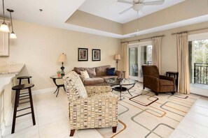 A spacious living room with a sofa, armchair, and a glass coffee table. There is a ceiling fan, a small table with a lamp, and a view of the outdoors through large windows.
