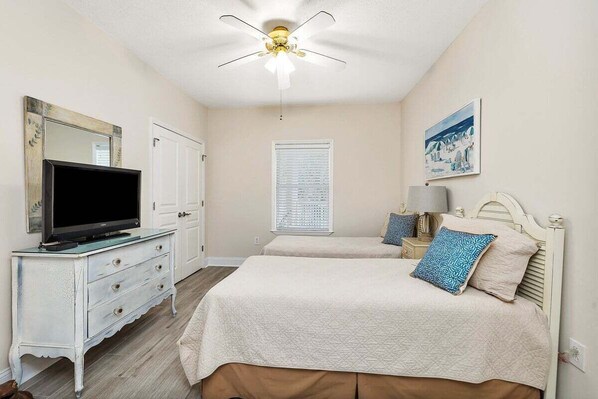 A bedroom with two beds, a television on a dresser, a ceiling fan, a window with blinds, and a painting on the wall. - Oceanfront Condo with Direct Access to Courtyard! (Saint Simons Island)