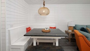 A dining area with a wooden table, white bench seating, a woven pendant light, and a teal sofa with orange cushions. - Oceanfront Condo with Balcony, Pool, Beach Access (Saint Simons Island)