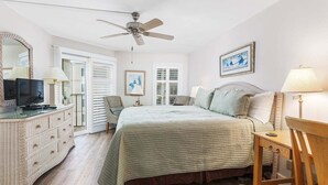 Un dormitorio con cama, televisión, cómoda, ventilador de techo y ventana.