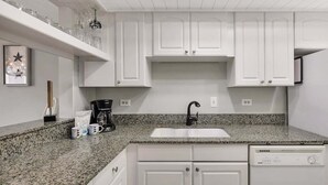 A kitchen with white cabinets, a granite countertop, a coffee maker, and a dishwasher. - Beachfront Condominium (Saint Simons Island)