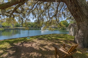 A wooden bench under a large tree by a calm lake with a fountain, surrounded by a well-maintained lawn and a small shed.