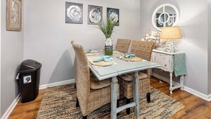 Un comedor con una mesa dispuesta para cuatro, rodeada de sillas de mimbre, un florero con plantas y una lámpara sobre una mesita.