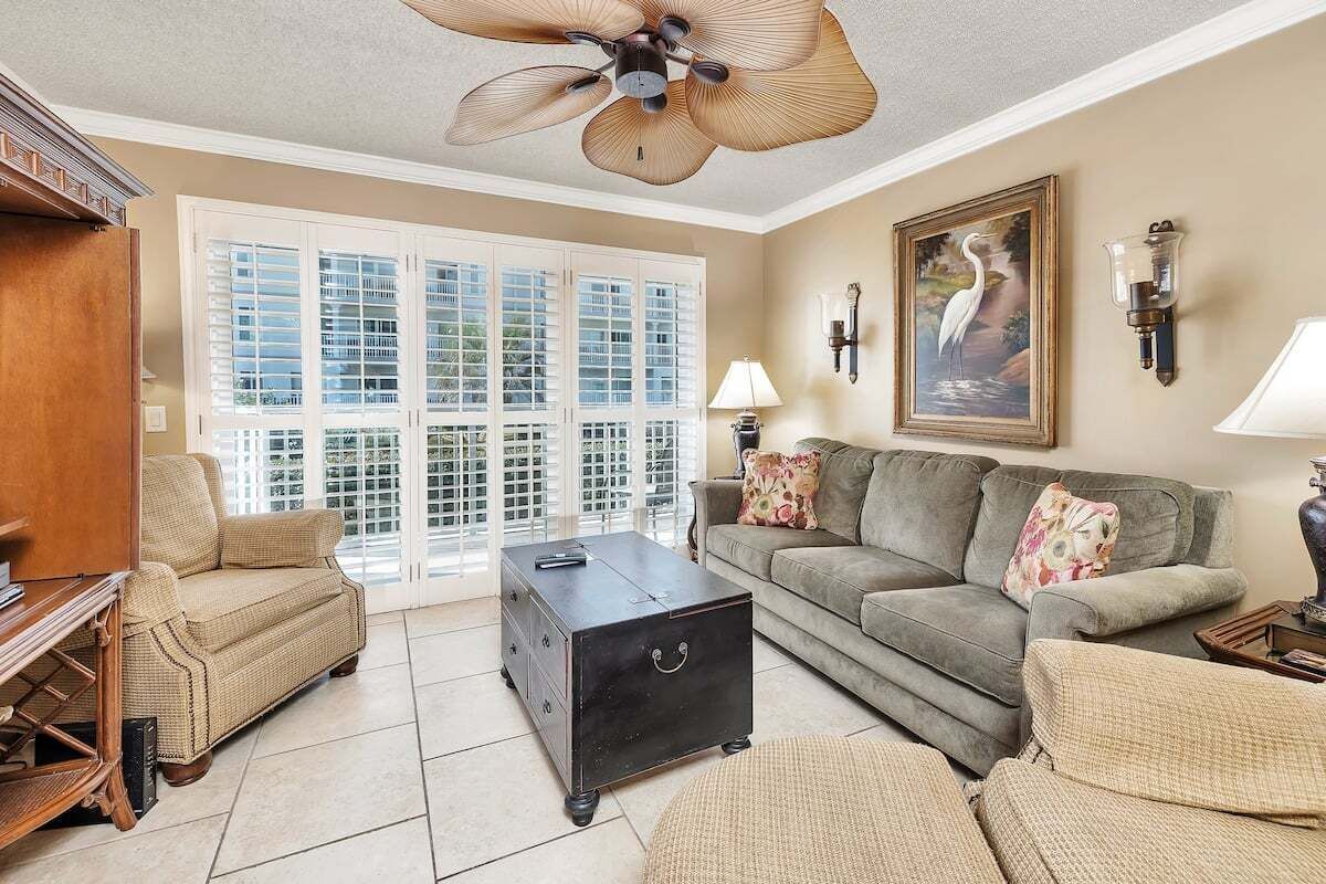 Ein Wohnzimmer mit beiger Couch, einem dunkelholzigen Couchtisch, einem Korbstuhl, einem Deckenventilator und einem großen Fenster mit Fensterläden.
