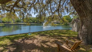 A wooden bench under a large tree by a calm lake with a fountain, surrounded by a well-maintained lawn and a small shed. - Ocean Walk Condo L14 (Saint Simons Island)