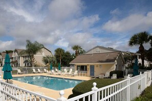 A swimming pool with lounge chairs and umbrellas, surrounded by a white fence and palm trees. - Ocean Walk Condo L14 (Saint Simons Island)