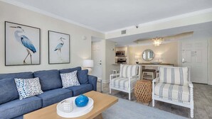 Ein Wohnzimmer mit einem blauen Sofa, einem Holztisch und einem gestreiften Sessel. An der Wand hängen zwei Bilder von Vögeln in Rahmen.