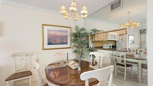 A dining area with a round wooden table, white chairs, a chandelier, and a large mirror reflecting the kitchen. - St Simons Island Spacious and Beachfront Condo! (Saint Simons Island)