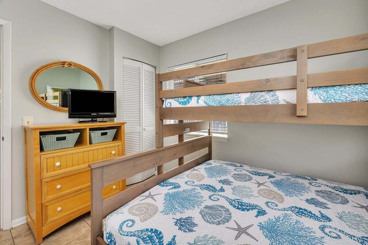 A bunk bed with a patterned bedspread, a wooden dresser with baskets, a television, and a round mirror on the wall.