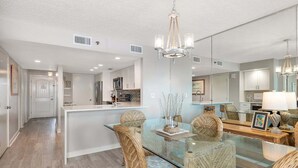 A modern kitchen with white cabinets, a central island, and a dining area with glass-top table and wicker chairs. - Oceanfront Condominium with Partial Ocean View! (Saint Simons Island)