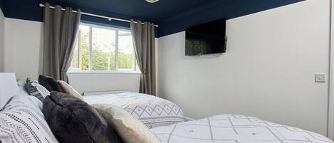 A bedroom with two beds, a window with curtains, a mounted TV, and a hanging light fixture.