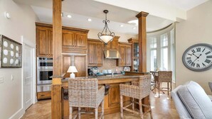 A spacious kitchen with wooden cabinets, a central island with bar stools, a large clock on the wall, and a dining area with wicker chairs. - Luxurious Complex, Ocean Views. Steps from Beach. (Saint Simons Island)