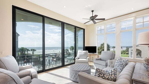 A spacious living room with a large sliding glass door leading to a balcony with a view of the ocean. There is a ceiling fan, a flat-screen TV, and a sofa with patterned cushions. - Luxurious Complex, Ocean Views. Steps from Beach. (Saint Simons Island)