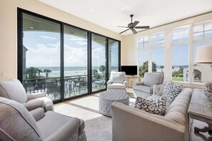 A spacious living room with a large sliding glass door leading to a balcony with a view of the ocean. There is a ceiling fan, a flat-screen TV, and a sofa with patterned cushions. - Luxurious Complex, Ocean Views. Steps from Beach. (Saint Simons Island)