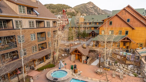 A multi-story building with a pool area, outdoor seating, and a mountain backdrop. - River Run Condo w/ Scenic Mountains Views; Steps from Gondola, Shops & Dining (Keystone)