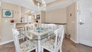 A dining area with a glass table, white chairs with floral upholstery, a chandelier, and a kitchen in the background. - Spacious Oceanfront Condominium on St Simons! (Saint Simons Island)