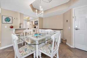 A dining area with a glass table, white chairs with floral upholstery, a chandelier, and a kitchen in the background.