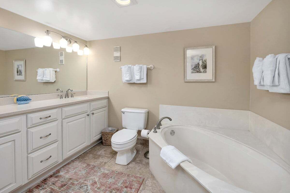 A bathroom with a white sink, toilet, and bathtub, beige walls, and a large mirror with lights.
