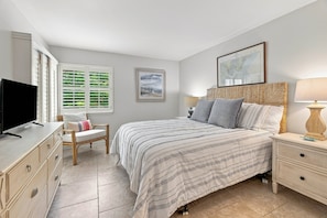 Une chambre à coucher comprenant un lit, une télévision, une chaise, une table de chevet et une fenêtre avec des stores.
