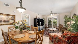 Uma zona de refeições com uma mesa e cadeiras de madeira, adjacente a uma sala de estar com um sofá e uma televisão.