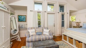 A cozy bedroom with a plaid sofa, a large mirror, and a bed with a white headboard. - Cottage Steps from the Beach with Community Pool (Saint Simons Island)