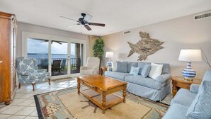 A living room with a sofa, armchairs, a coffee table, and a ceiling fan. There is a large window with a view outside. - First Floor Oceanfront Condominium in the Heart of (Saint Simons Island)