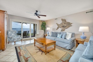 A living room with a sofa, armchairs, a coffee table, and a ceiling fan. There is a large window with a view outside.
